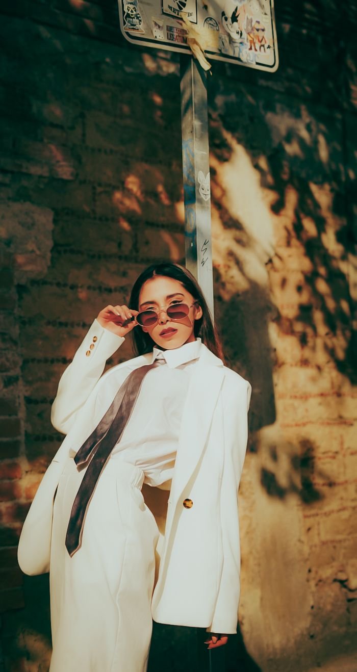 services-02 Fashionable woman in a white suit posing with sunglasses against a brick wall in warm sunlight.
