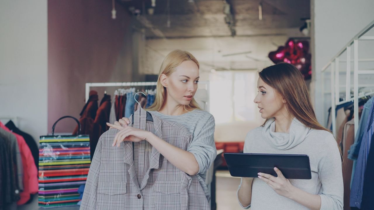 our-experience Two women in trendy boutique discussing clothing and style options, holding a plaid jacket.