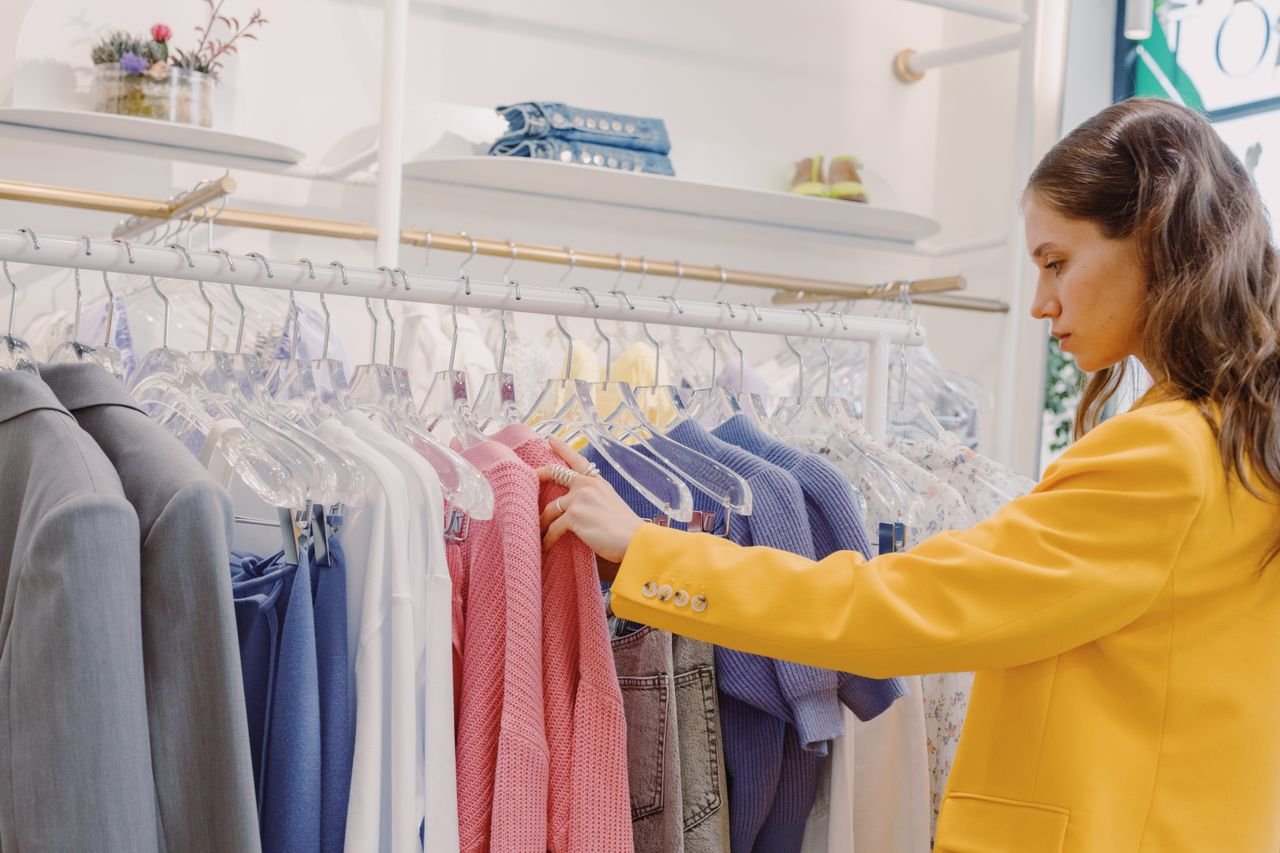 hero-img-02 Woman browsing clothes on a rack in a clothing store, wearing a stylish yellow blazer.