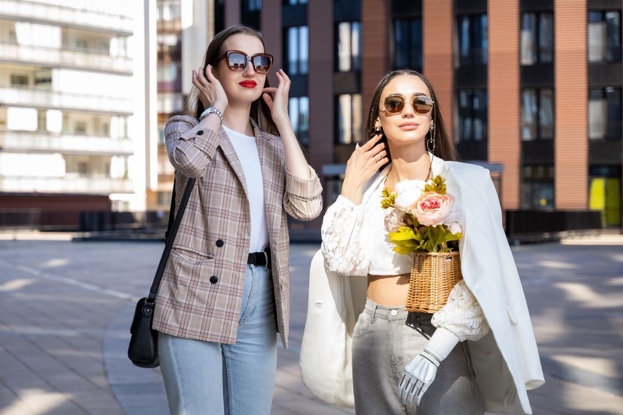 about-img Two stylish women enjoy a sunny day in the city wearing chic outfits and sunglasses.