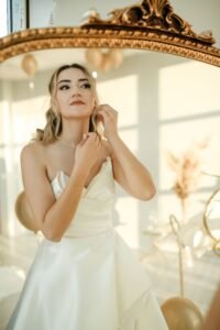 Bride trying on wedding earrings in front of a mirror with wedding dress in background